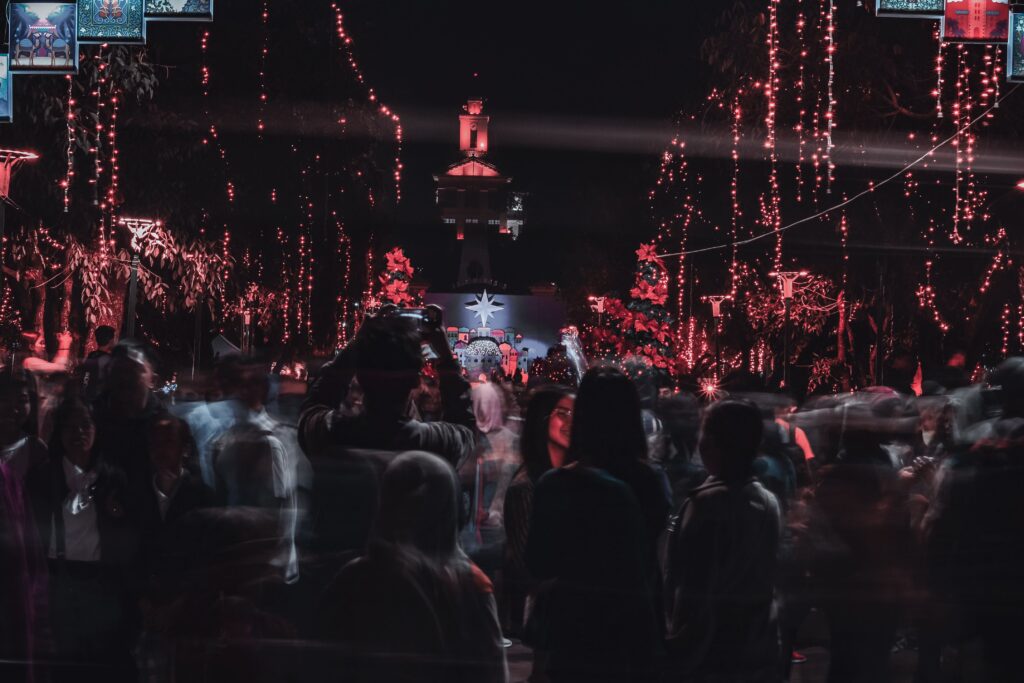 A lively crowd gathers at night in a decorated square with festive lights and a church backdrop.