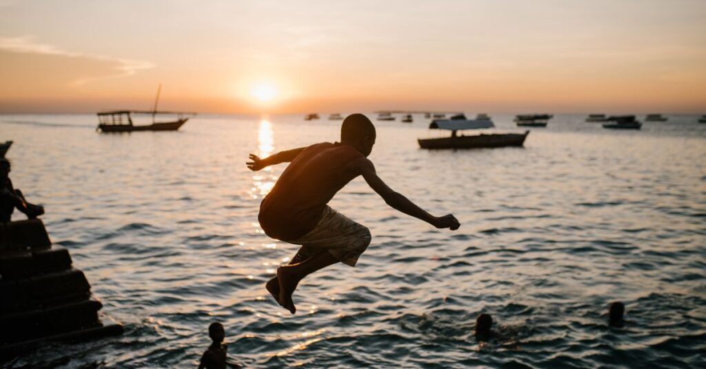 Summer 2026: How to Make It Count Back view of anonymous ethnic teen boy in moment of jumping in sea water at sunset
