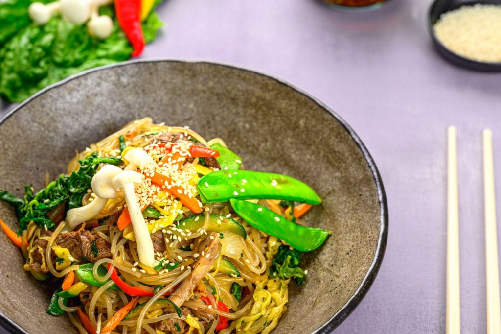 A vibrant bowl of Asian stir-fry with colorful vegetables, mushrooms, and sesame seeds served with chopsticks.