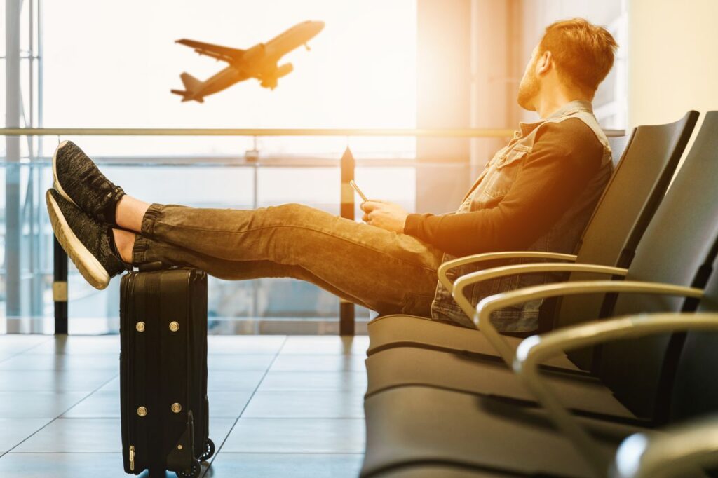 Side view full body of bearded man sitting on chair in waiting hall of airport and looking at flying aircraft behind window