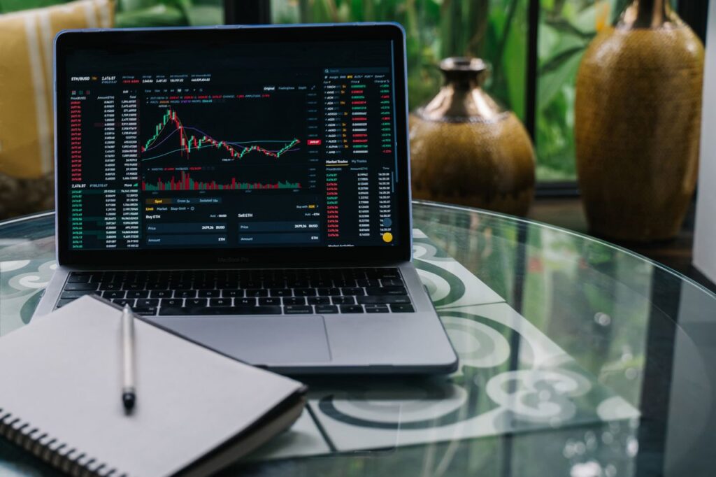 One Meeting Runs Your Whole Portfolio Now Laptop displaying cryptocurrency stocks and graphs on a glass table with a notepad.