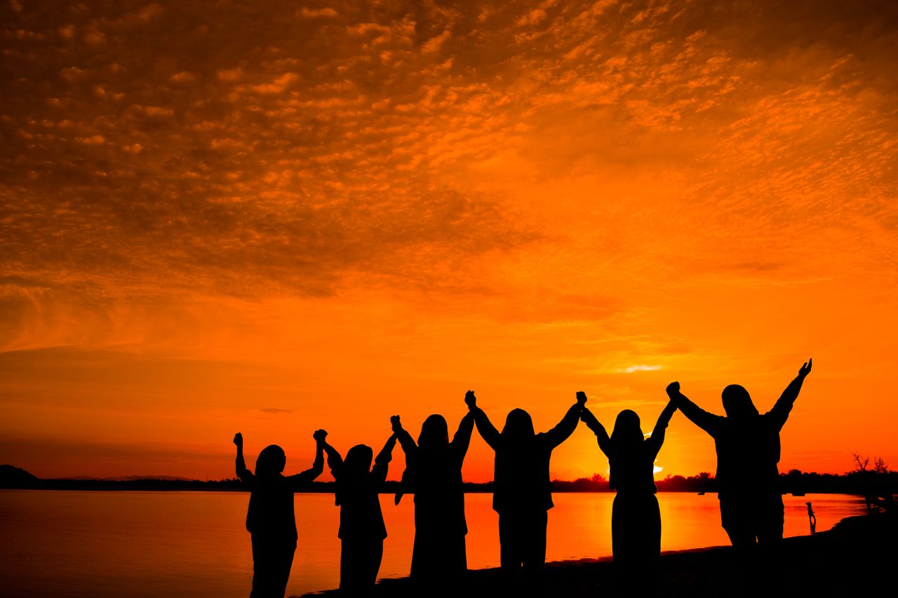Group of silhouetted people holding hands against a vibrant orange sunset by the lakeshore.