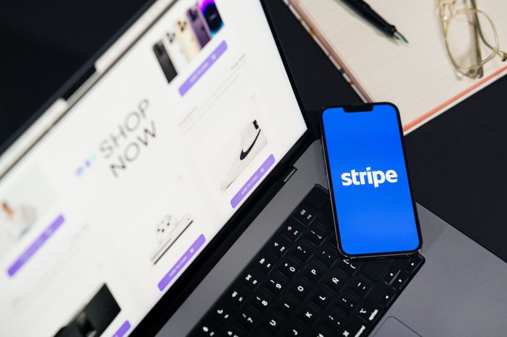 Stripe, PayPal and the Quiet Dollar Race Mobile phone displaying Stripe app on a laptop with an eCommerce site open, symbolizing online shopping.