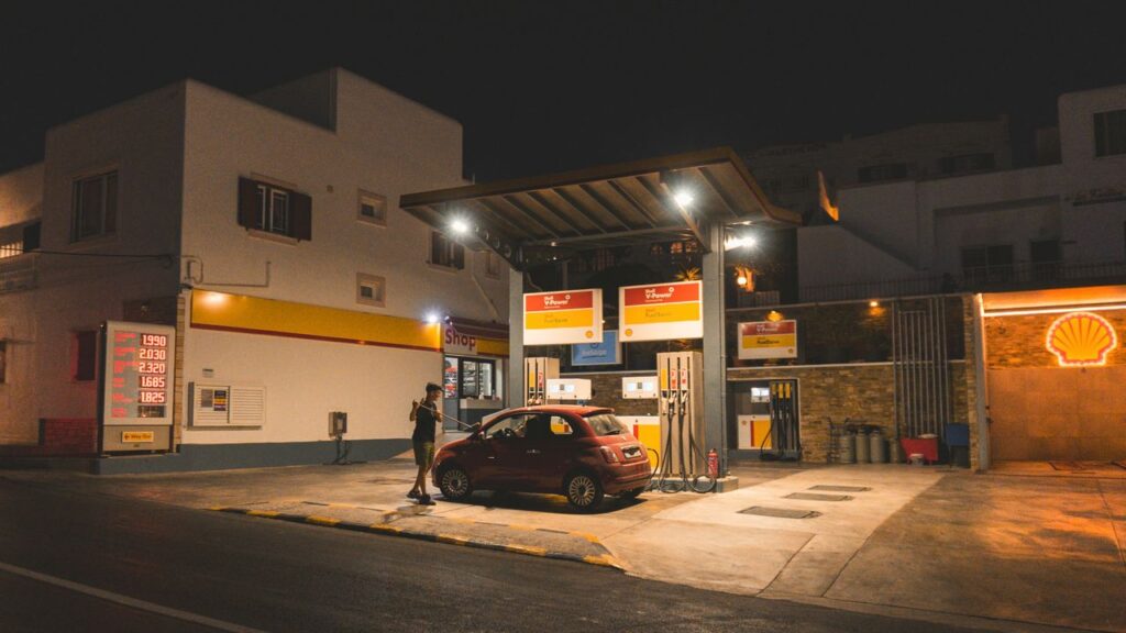 The War You're Not Fighting And Why It's Already at Your Petrol Pump A person refuels a red car at a gas station at night, illuminated signs and lights.