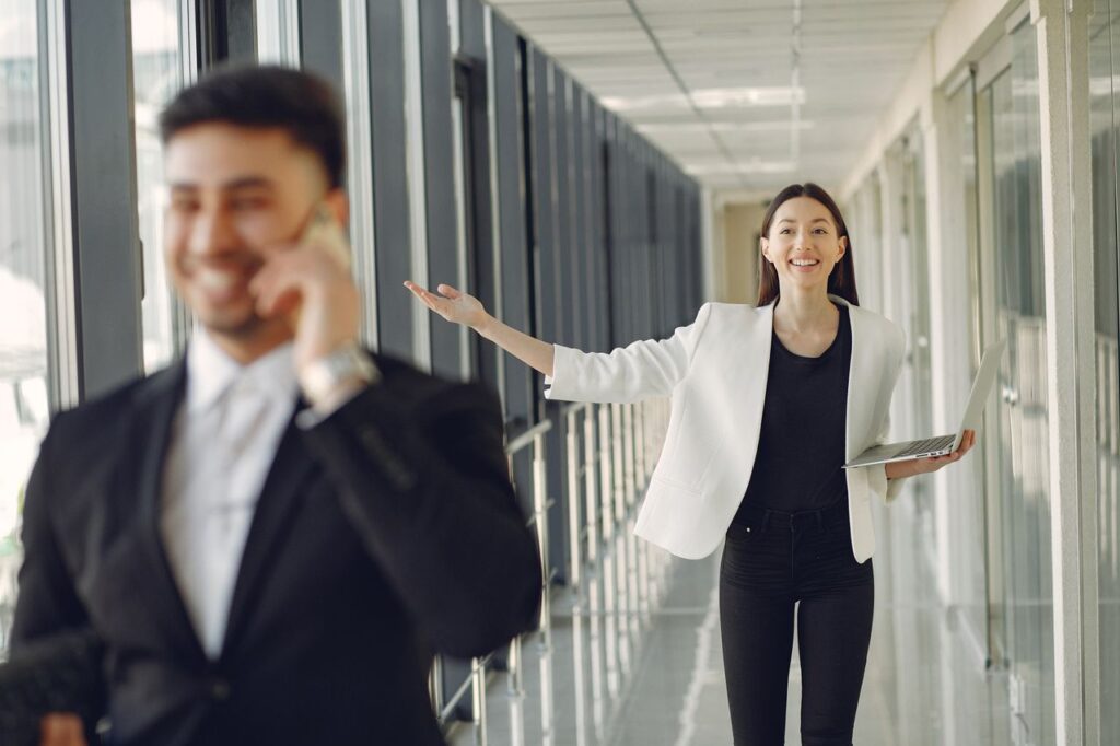 Positive female in formal outfit standing with laptop in modern office and looking at male employee talking on mobile phone