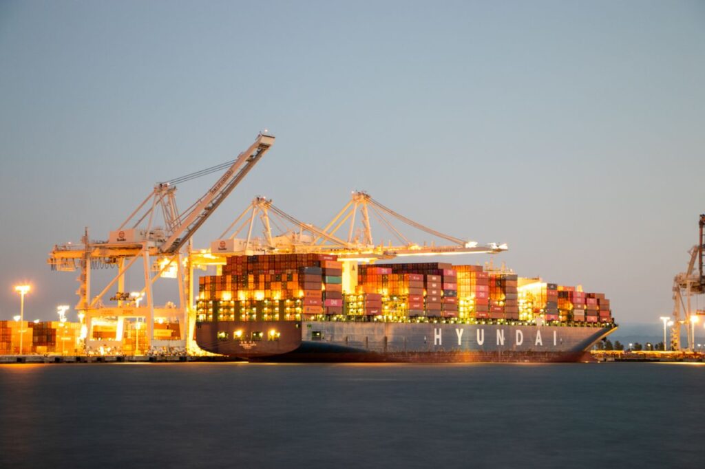 The Tariffs Landed This Morning. Now What? Container ship docked at port under cranes during twilight, illuminated against the evening sky.