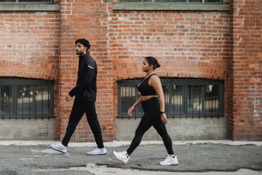 The Most Underrated Exercise Is Just Walking A man and woman walking briskly in sportswear against a brick wall in the city.
