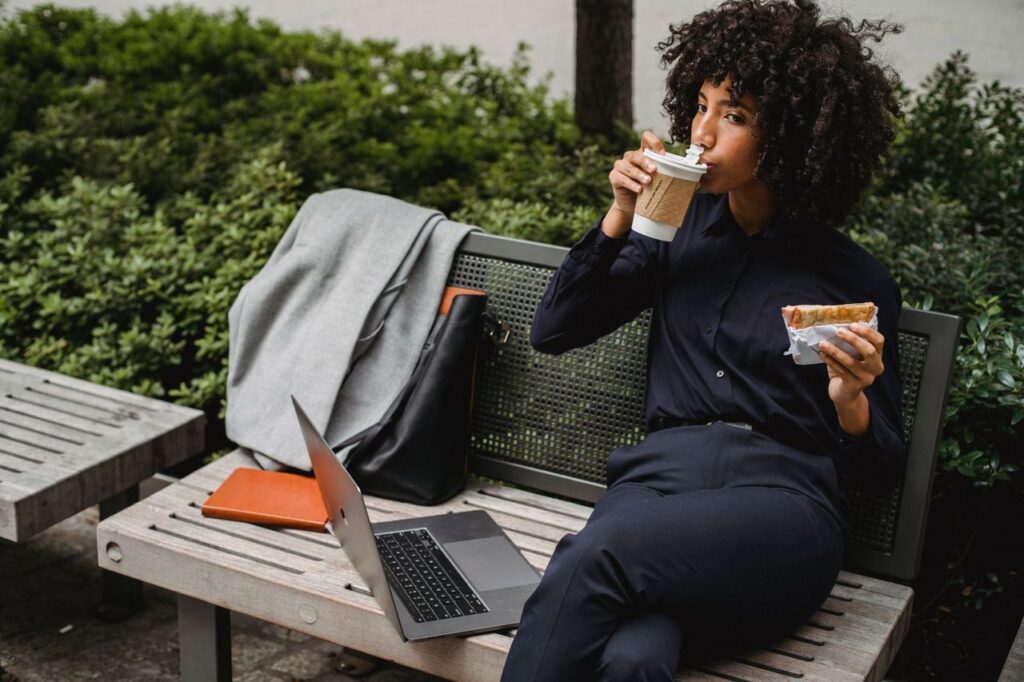The Long Lunch Is a Power Move Adult woman sipping coffee and eating a sandwich while working outdoors on a bench.