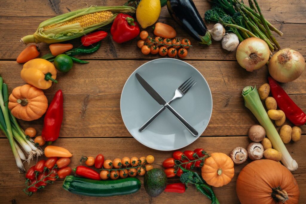 The Eating Window Is Not the Magic Top view of fresh vegetables creatively arranged around an empty plate on a wooden table.