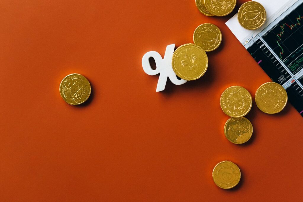 Three Central Banks, One Lesson for Your Money Gold coins scattered with a stock market graph and a percentage symbol on an orange background.