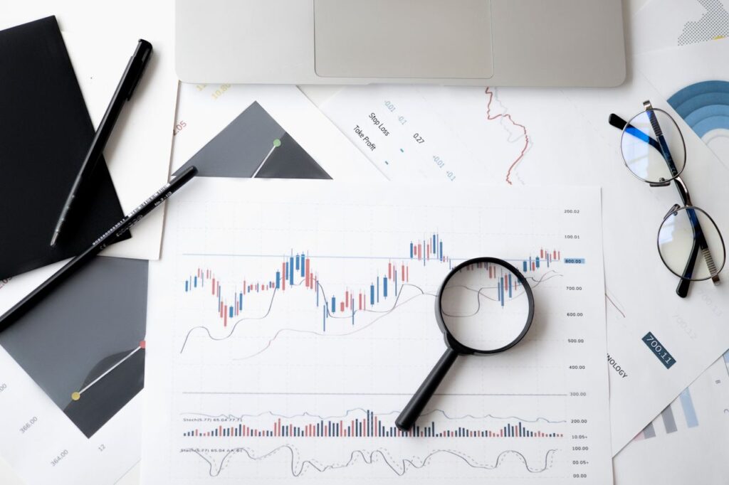 Finance Was Built to Sound Harder Than It Is Flat lay of stock market analysis documents with magnifying glass, pens, and glasses.