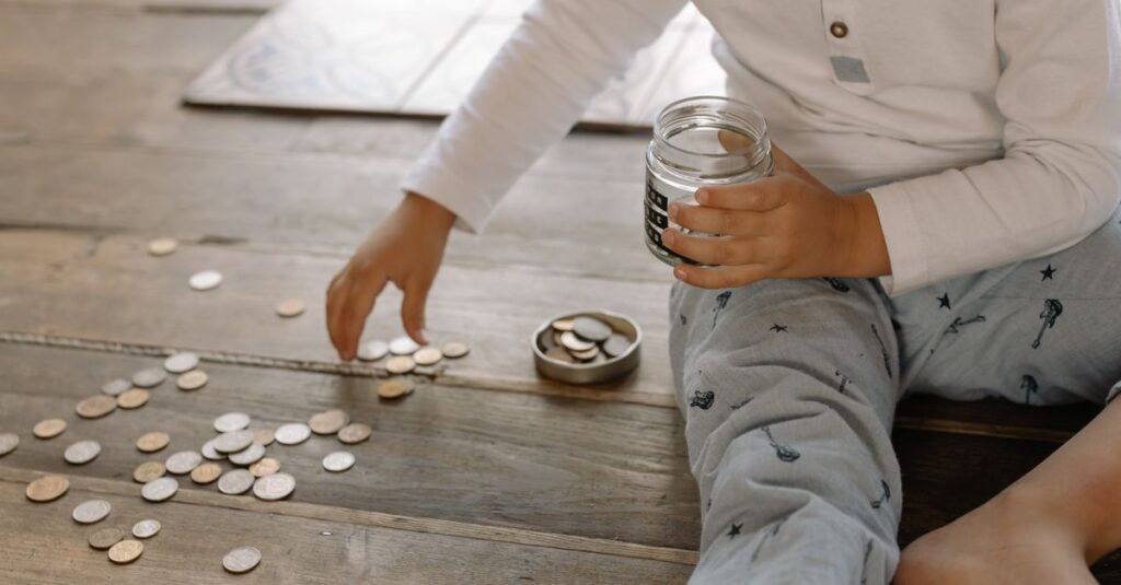 How to Save Money: 10 Changes That Actually Move the Needle (2026) A young child collects coins into a jar on a wooden floor, symbolizing savings.