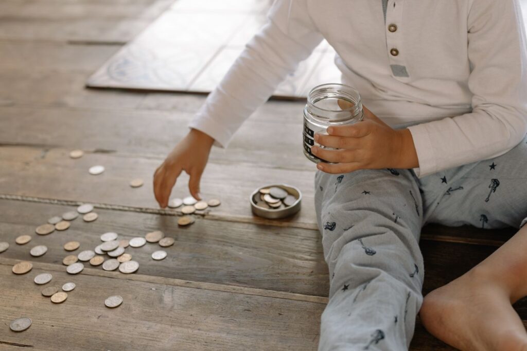When Boring Money Finally Pays A young child collects coins into a jar on a wooden floor, symbolizing savings.