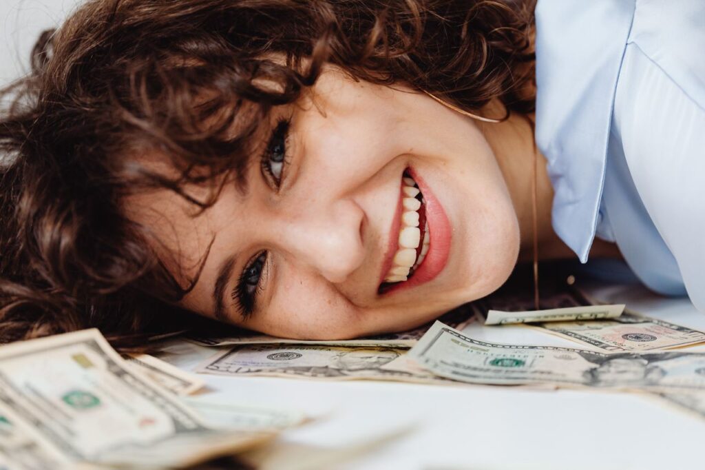 The Bravest Word in Finance Is "Enough" A joyful woman with curly hair lies on a pile of dollar bills, smiling brightly.
