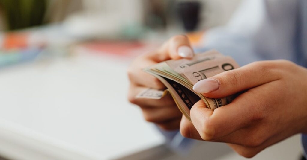 The Bravest Word in Finance Is "Enough" Close-up shot of hands carefully counting US dollar bills indoors at a desk.