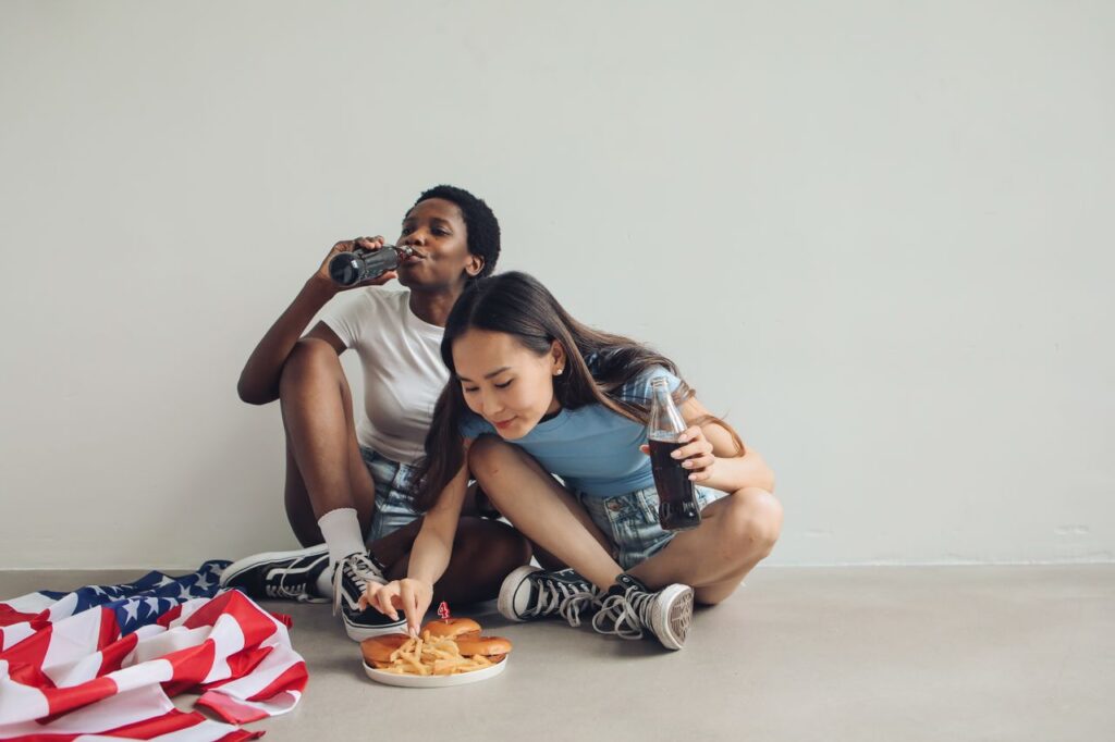Two friends enjoying snacks and soda indoors beside an American flag.