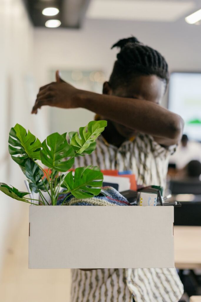 When the Job Goes A man in an office carrying a white box filled with personal items and a plant, covering his face.