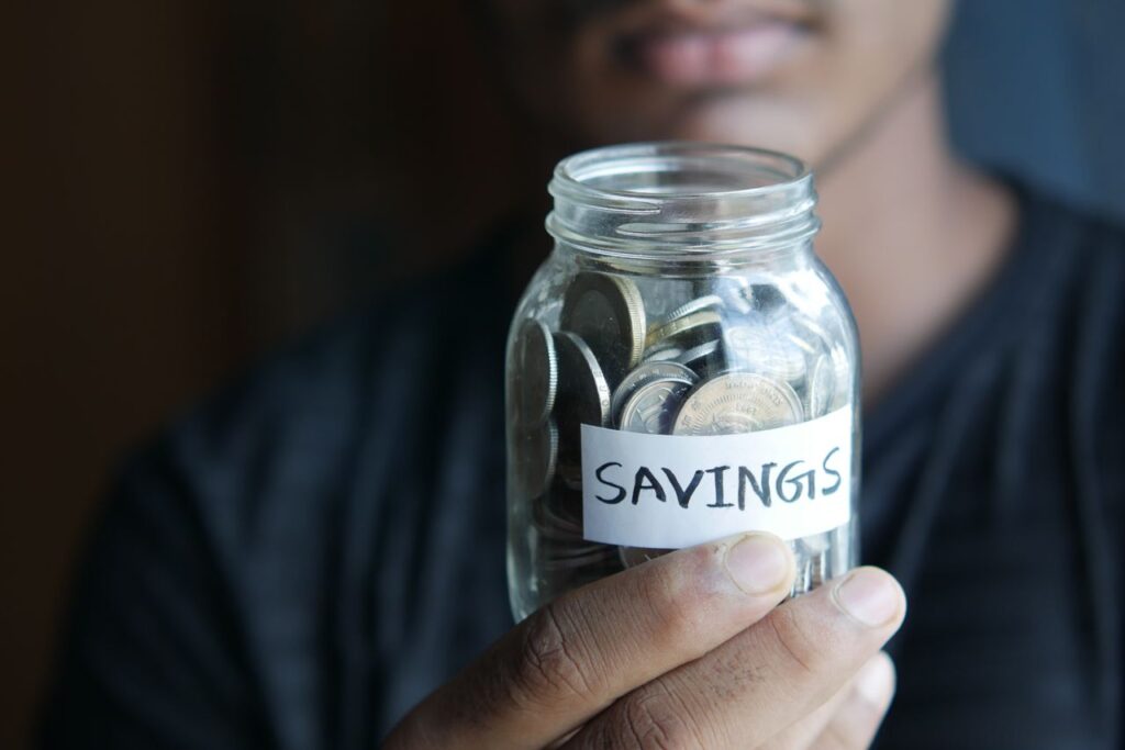 How to Save Money: 10 Changes That Actually Move the Needle (2026) A close-up image of a person's hand holding a jar full of coins labeled 'Savings'.