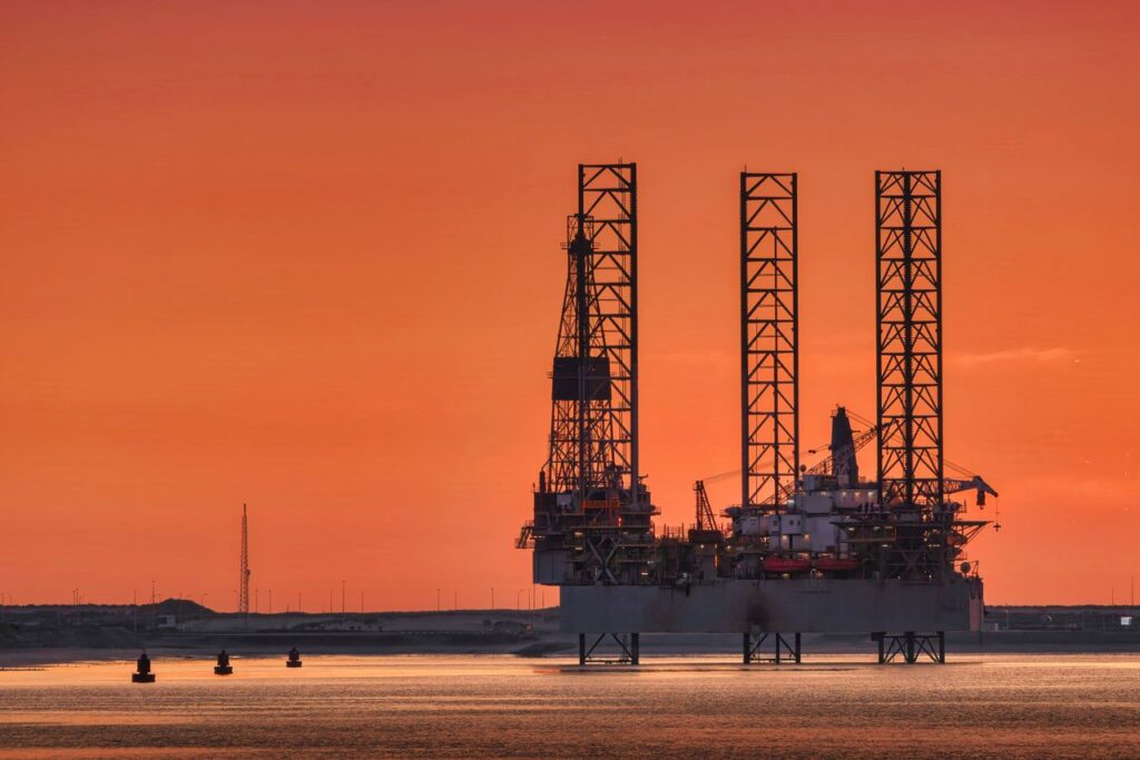 Oil Below Eighty. What It Means at the Pump A striking view of an oil rig silhouetted against a fiery sunset in Rotterdam harbor.