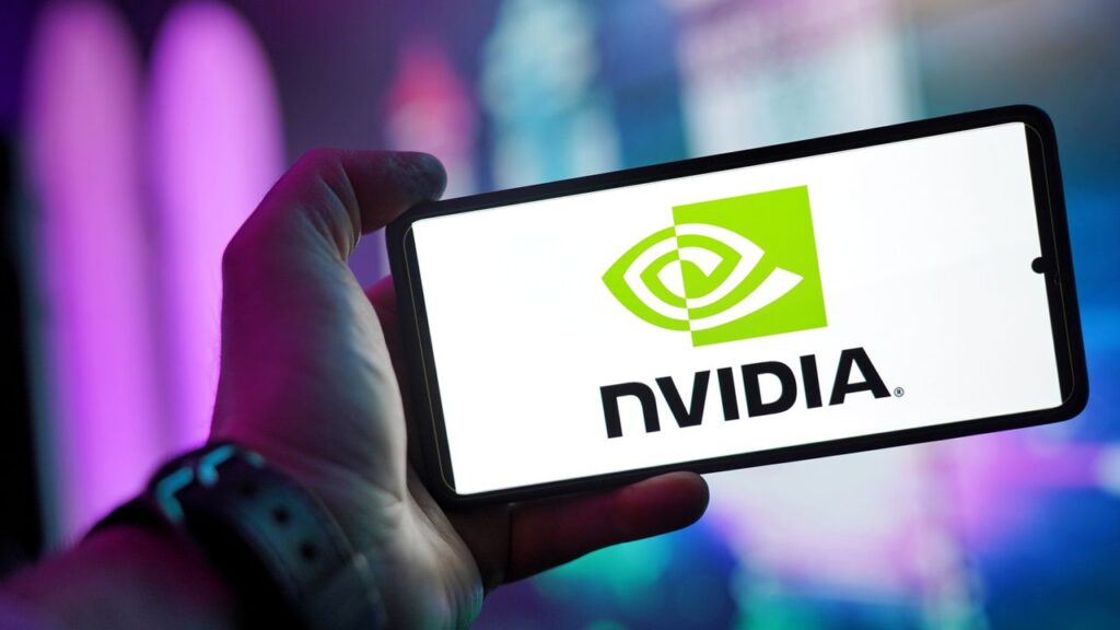 Nvidia Beat Every Number and Still Got Punished Close-up of a hand holding a smartphone showing the NVIDIA logo on screen with a blurred background.