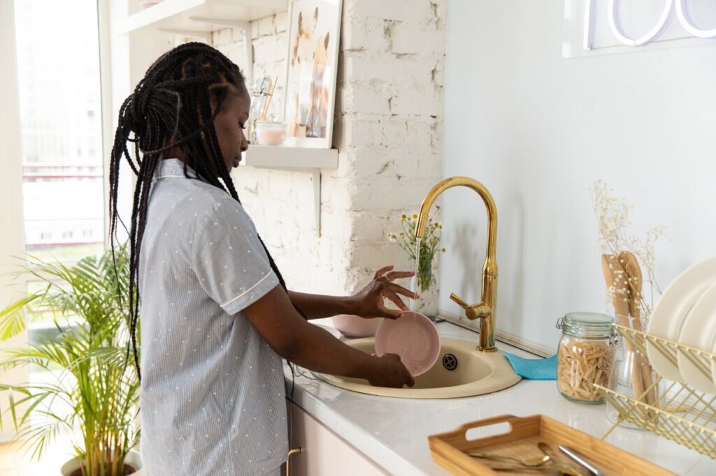 The Tap I've Been Ignoring Since March An adult woman washing dishes in a bright, modern kitchen setting.