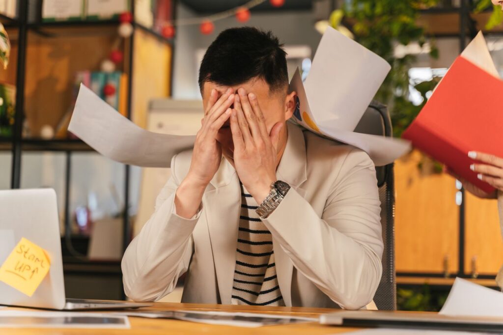 The Business Doesn't Care That You're Tired Stressed businessman overwhelmed by paperwork in office environment, demonstrating burnout.