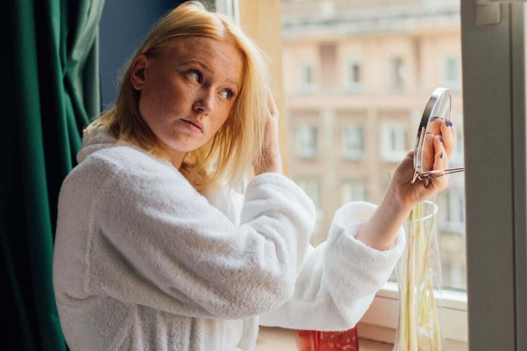 I Got More Attractive by Doing Less to My Face A woman in a white bathrobe looking in a mirror by the window.