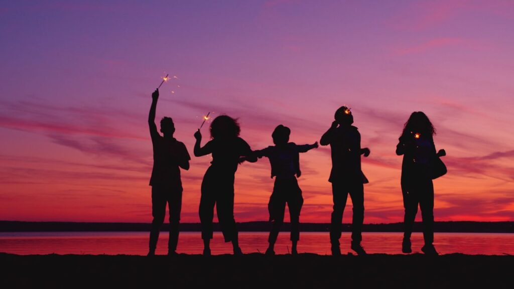 You Earn Well. So Why Doesn't Life Feel Freer? Five people holding sparklers in silhouette against a vibrant sunset sky by the water.