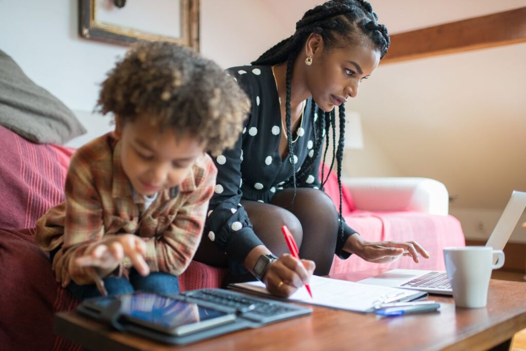 Walking Through Treacle, and Why I Keep Walking A mother multitasking at home, working on a laptop while her child plays on a tablet nearby.