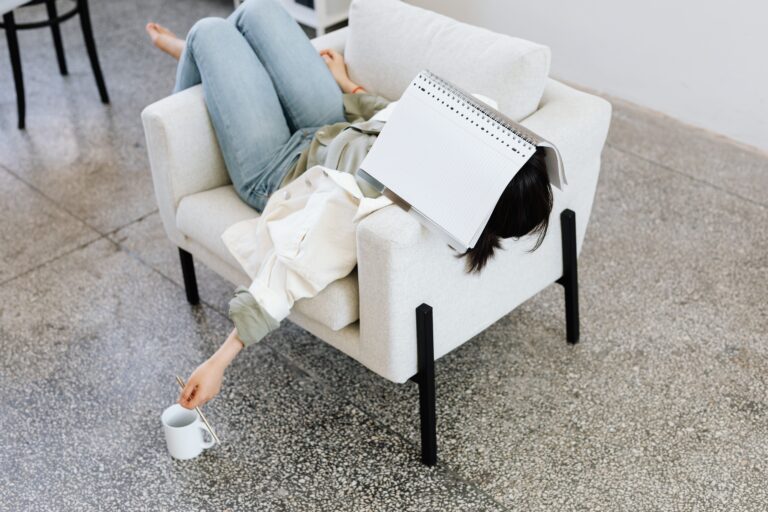 Woman lying on an armchair with a book on face, holding a coffee. Casual and relaxing atmosphere.