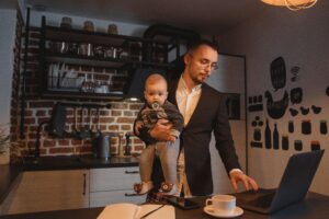 Articles Father working on a laptop while holding his baby in a kitchen setting.