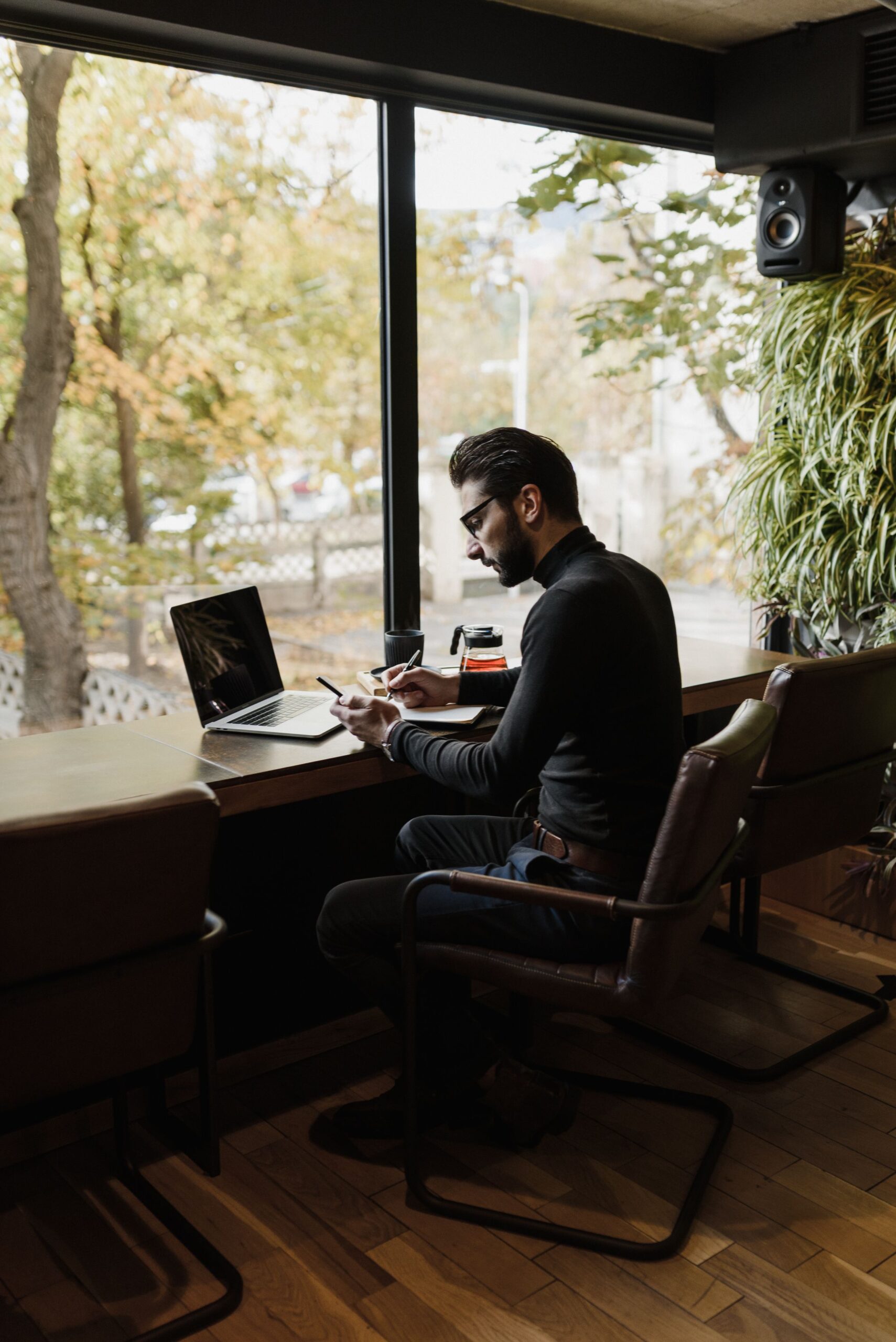 Man working on a laptop near a large window in a cozy cafe, focusing on tasks with autumn view outside.
