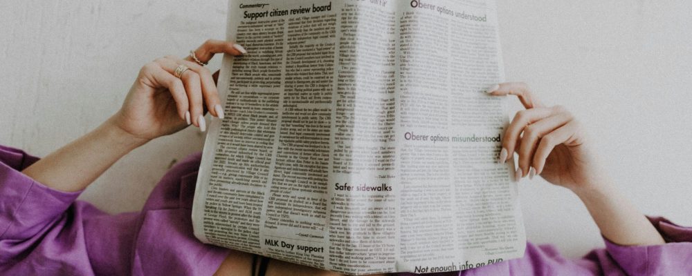 A woman in a violet jacket holds a newspaper, obscuring her face against a light background.